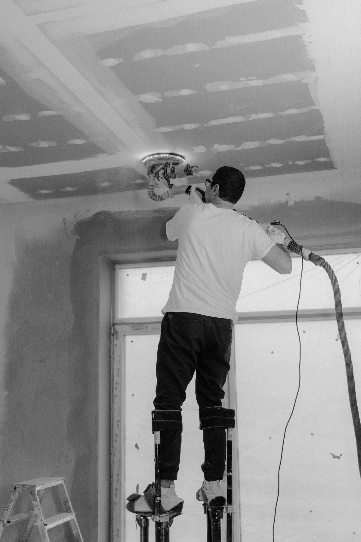 Tradesman on drywall stilts running a dust-collecting sander across a taped ceiling, shown in black and white