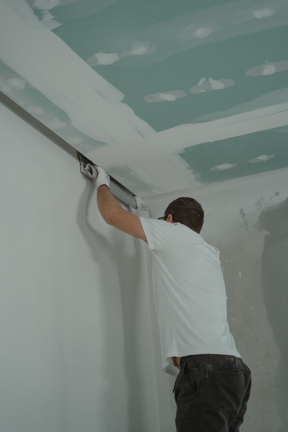 Worker finishing the joint where new ceiling drywall meets the top of a wall with a long taping knife