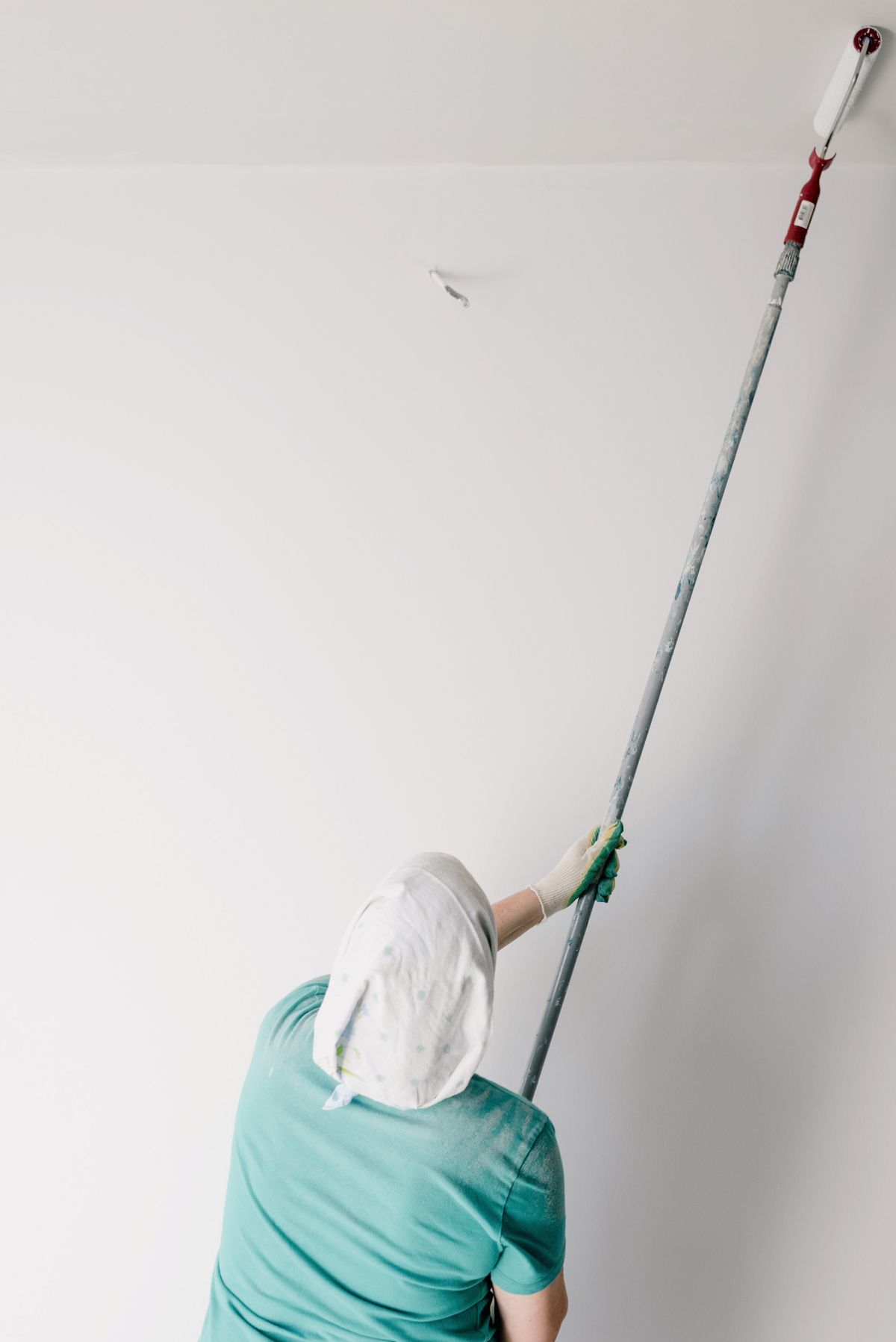 Painter in a head covering rolling white paint onto a ceiling using a roller on a long extension pole